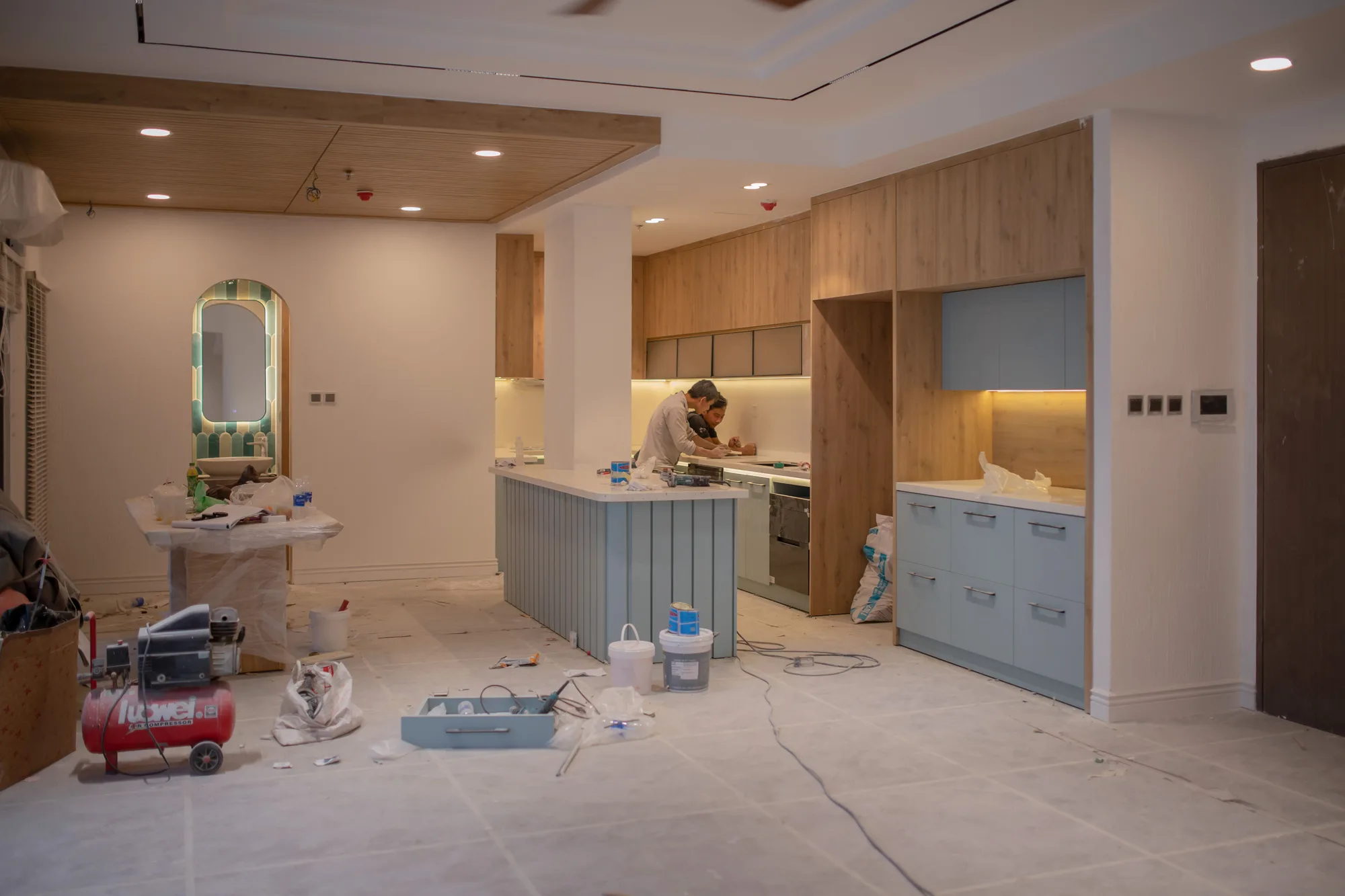 Cuisine en cours de rénovation : deux artisans au travail sur l’îlot, meubles en bois et bleu clair, sol protégé, compresseur et outils au premier plan.